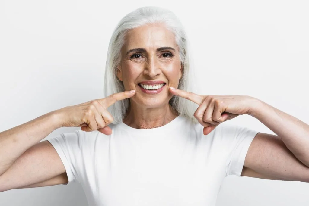 Mulher idosa de cabelos brancos apontando para o sorriso, vestindo uma camiseta branca, com fundo branco minimalista.